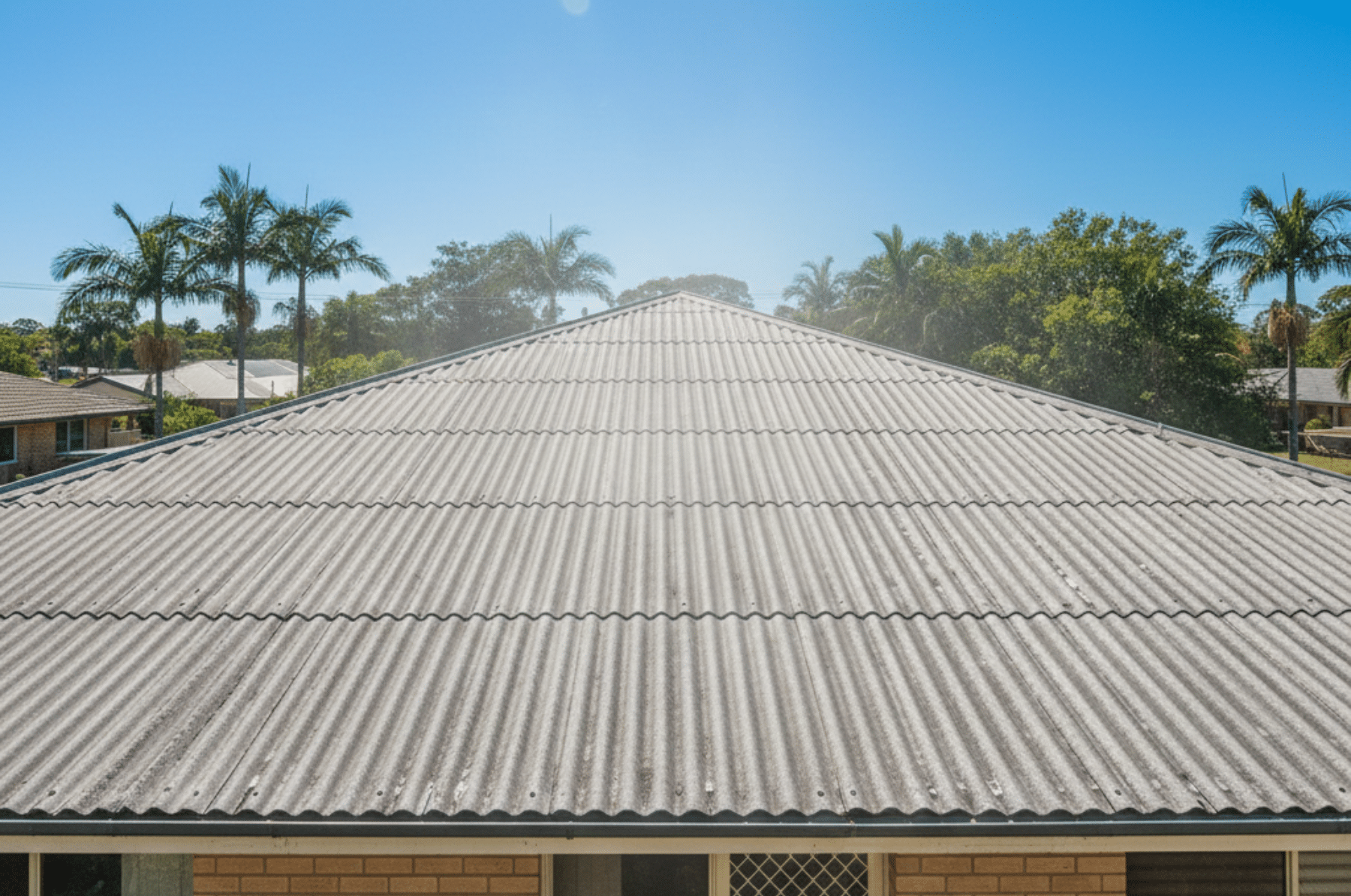 Close-up view of a corrugated metal roof on a single-story brick house under a clear blue sky, with palm trees in the background.